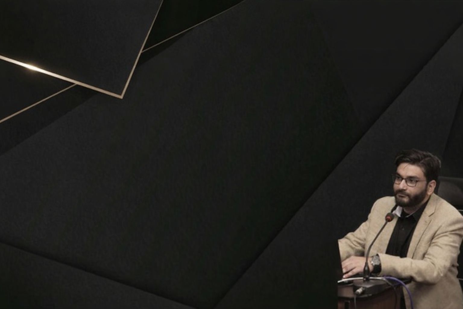 Mies silmälaseissa ja beigessä takissa puhumassa mikrofooniin, tummaa taustaa vasten.